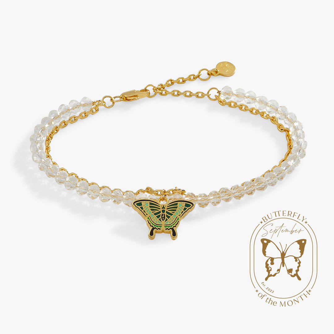 A double-strand beaded bracelet with clear faceted beads, gold-tone chains, and a central gold butterfly charm with green enamel detailing, shown on a light gray background with a Butterfly of the Month badge.