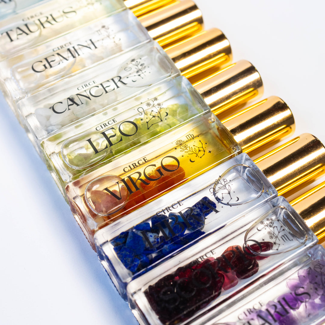 A close-up diagonal arrangement of small clear glass vials with gold caps, each labeled with a zodiac name such as Taurus, Gemini, Cancer, Leo, Virgo, and Pisces. The vials contain colorful translucent liquid with embedded gem-like pieces in blue, purple, red, orange, and green.