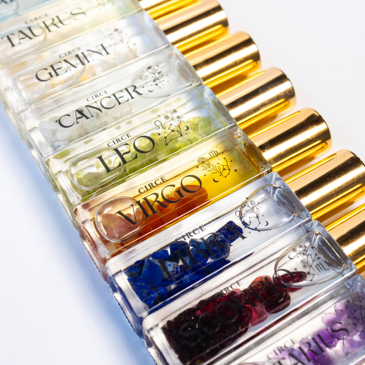 A close-up diagonal arrangement of small clear glass vials with gold caps, each labeled with a zodiac name such as Taurus, Gemini, Cancer, Leo, Virgo, and Pisces. The vials contain colorful translucent liquid with embedded gem-like pieces in blue, purple, red, orange, and green.
