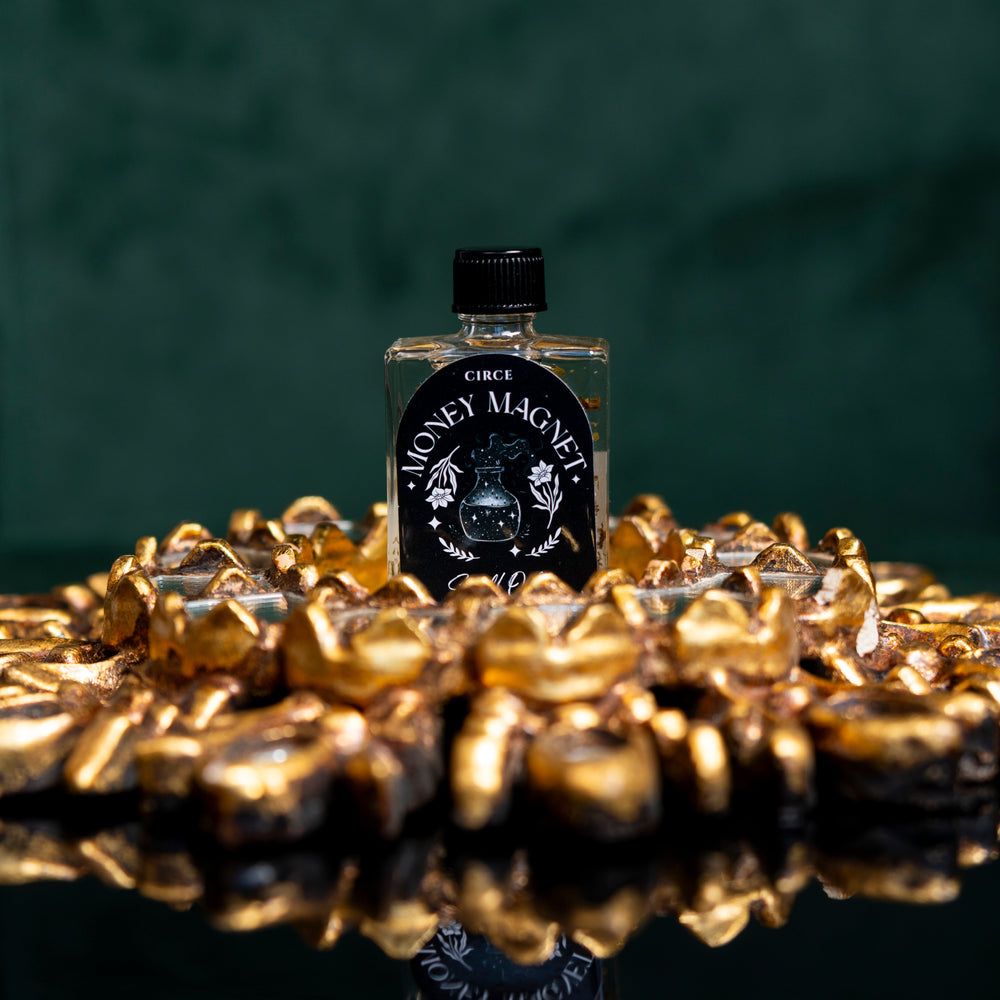 Small bottle labeled 'Money Magic' surrounded by gold coins on a reflective surface with a dark background