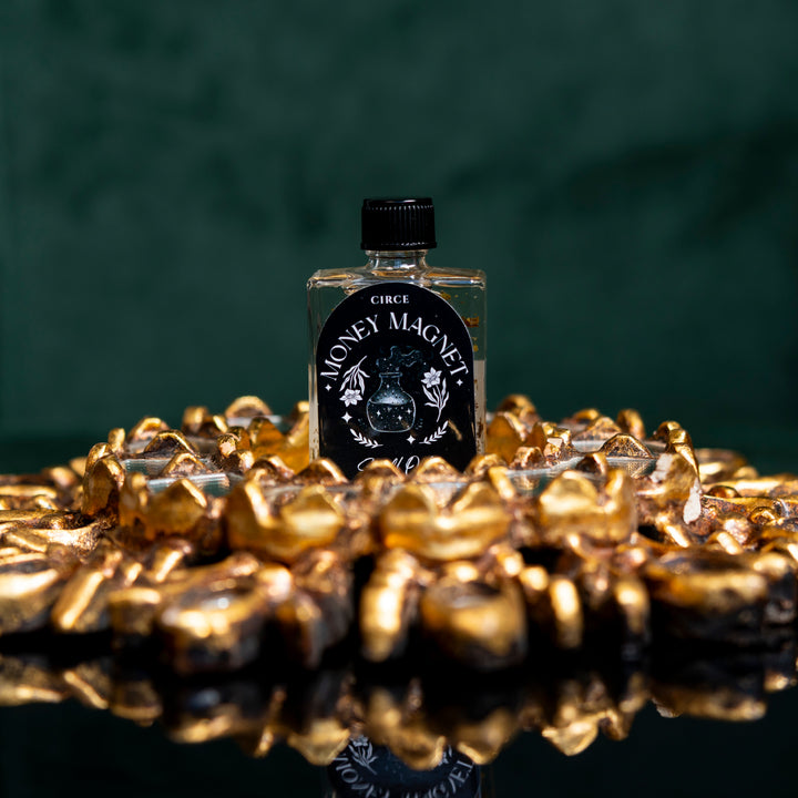 Small bottle labeled 'Money Magic' surrounded by gold coins on a reflective surface with a dark background