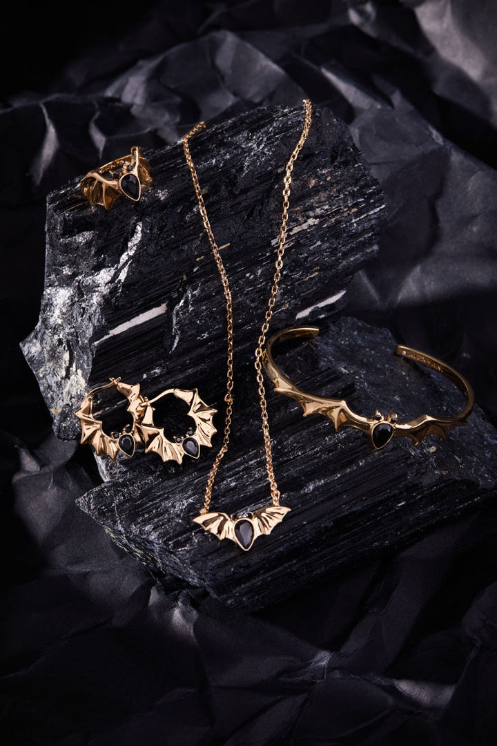 Gold jewelry bat set including necklace, earrings, and bracelet on black stone and fabric background