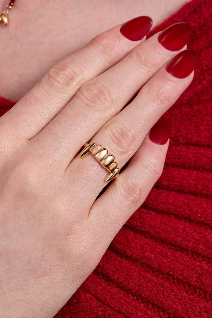 Hand wearing a gold vampire ring with red nail polish against a red background