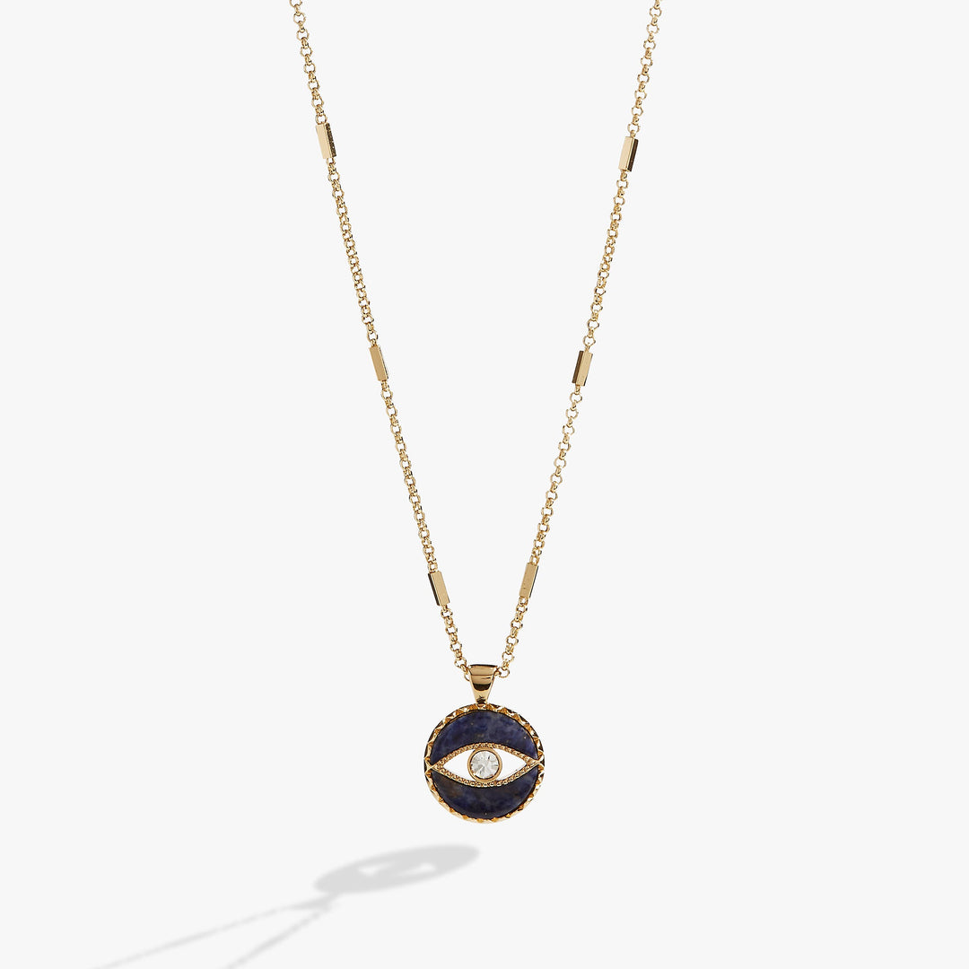 A gold chain necklace featuring a circular pendant with a dark navy blue enamel background and a central gold eye motif with a clear stone, shown against a light gray background.