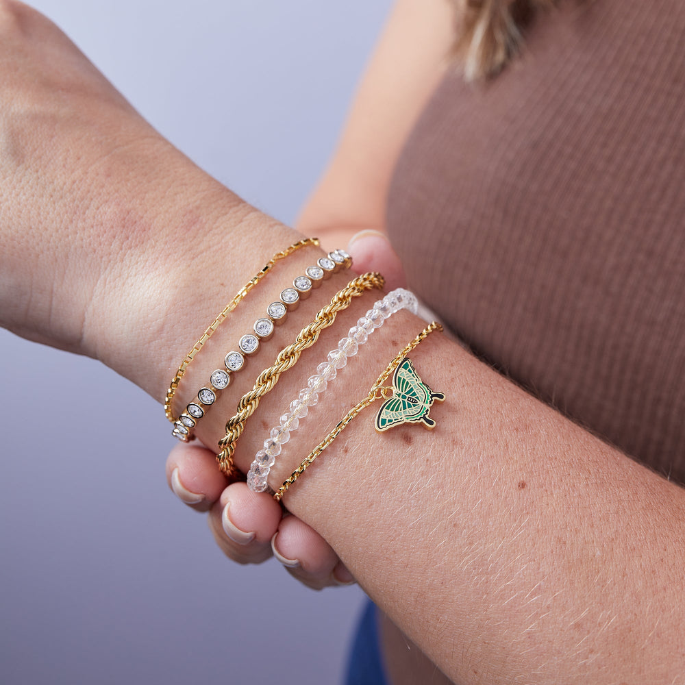 Close-up of a wrist wearing multiple gold and pearl bracelets with a green butterfly charm against a blurred background.