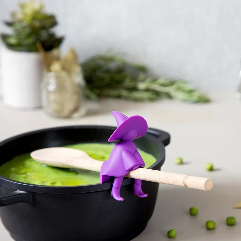 Purple witch figurine with a wooden broom stirring green soup in a black pot.