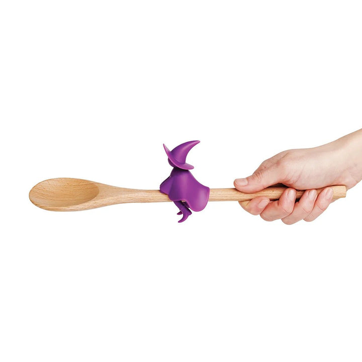 Hand holding a wooden spoon with a purple witch-shaped handle on a white background