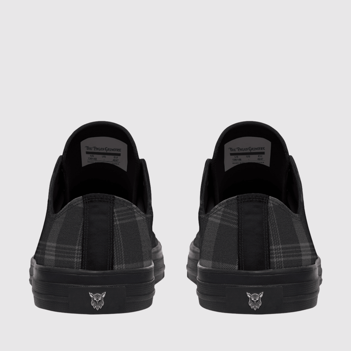 A pair of black slip-on sneakers shown from the back, featuring a black and gray plaid pattern on the sides and a small silver wolf head logo on the black rubber soles.