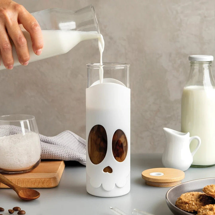 Person pouring milk into a glass with a skull design on a kitchen counter.