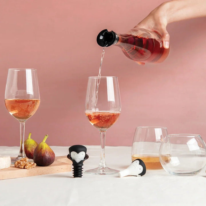 A hand pours red wine from a glass bottle into two wine glasses on a table, with figs, cheese, and a small black-and-white heart-shaped corkscrew or stopper nearby against a pink background.
