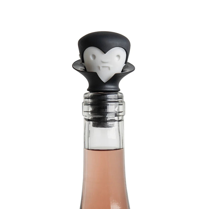 A close-up of a clear glass bottle with a black and white vampire-shaped stopper on a white background.