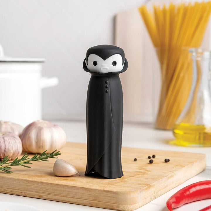 A black pepper mill shaped like a cartoon vampire in a tuxedo stands on a wooden cutting board in a kitchen setting, with garlic, rosemary, peppercorns, spaghetti, and a red chili pepper nearby.