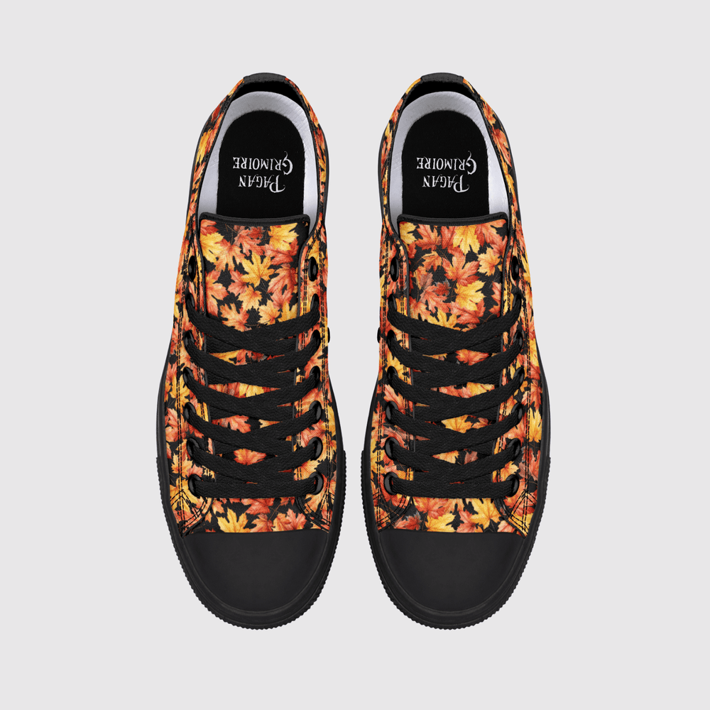 A pair of low-top canvas sneakers shown from a top-down view on a light gray background. The uppers feature a vibrant autumn leaf pattern in shades of orange, yellow, and red, with black laces, black rubber toe caps, and black inner soles.