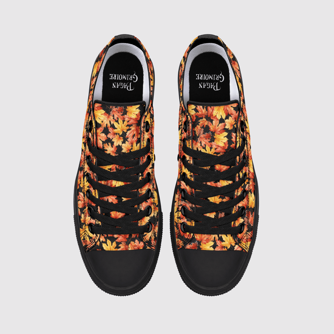A pair of low-top canvas sneakers shown from a top-down view on a light gray background. The uppers feature a vibrant autumn leaf pattern in shades of orange, yellow, and red, with black laces, black rubber toe caps, and black inner soles.