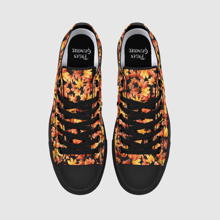 A pair of low-top canvas sneakers shown from a top-down view on a light gray background. The uppers feature a vibrant autumn leaf pattern in shades of orange, yellow, and red, with black laces, black rubber toe caps, and black inner soles.