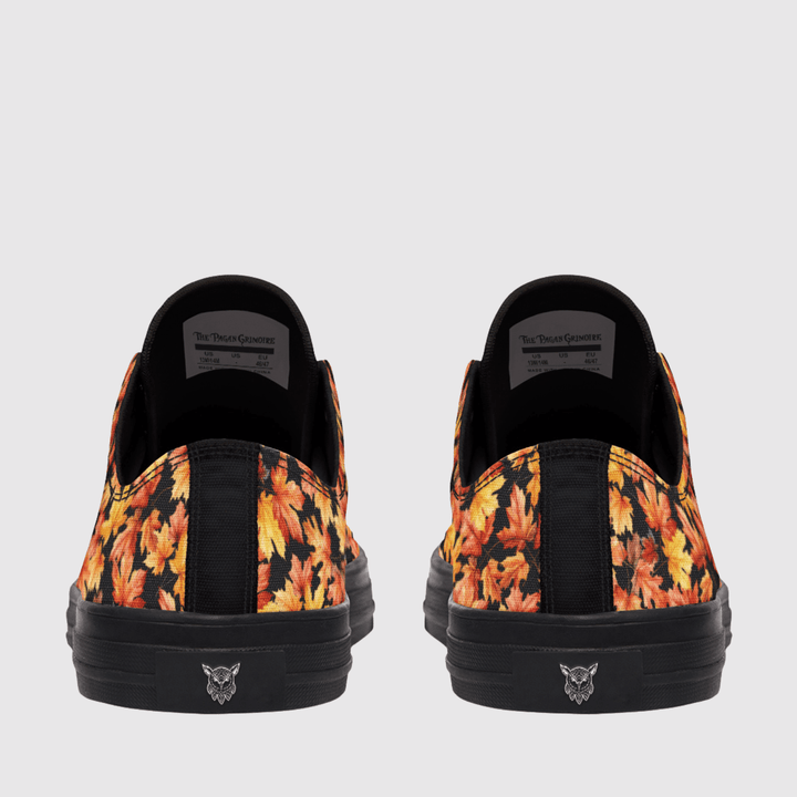A pair of low-top slip-on sneakers shown from the back, featuring a black base with a vibrant orange, yellow, and red autumn leaf pattern on the sides and a black sole with a small logo.