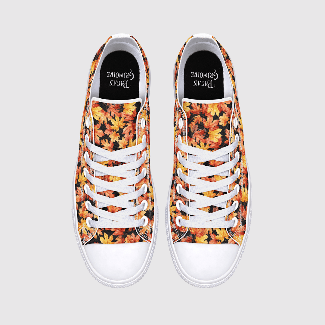 A pair of low-top canvas sneakers shown from a top-down view on a light gray background. The uppers feature a dense autumn leaf pattern in orange, yellow, red, and brown, with white laces, white rubber toe caps, and a black insole.