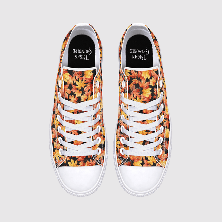 A pair of low-top canvas sneakers shown from a top-down view on a light gray background. The uppers feature a dense autumn leaf pattern in orange, yellow, red, and brown, with white laces, white rubber toe caps, and a black insole.