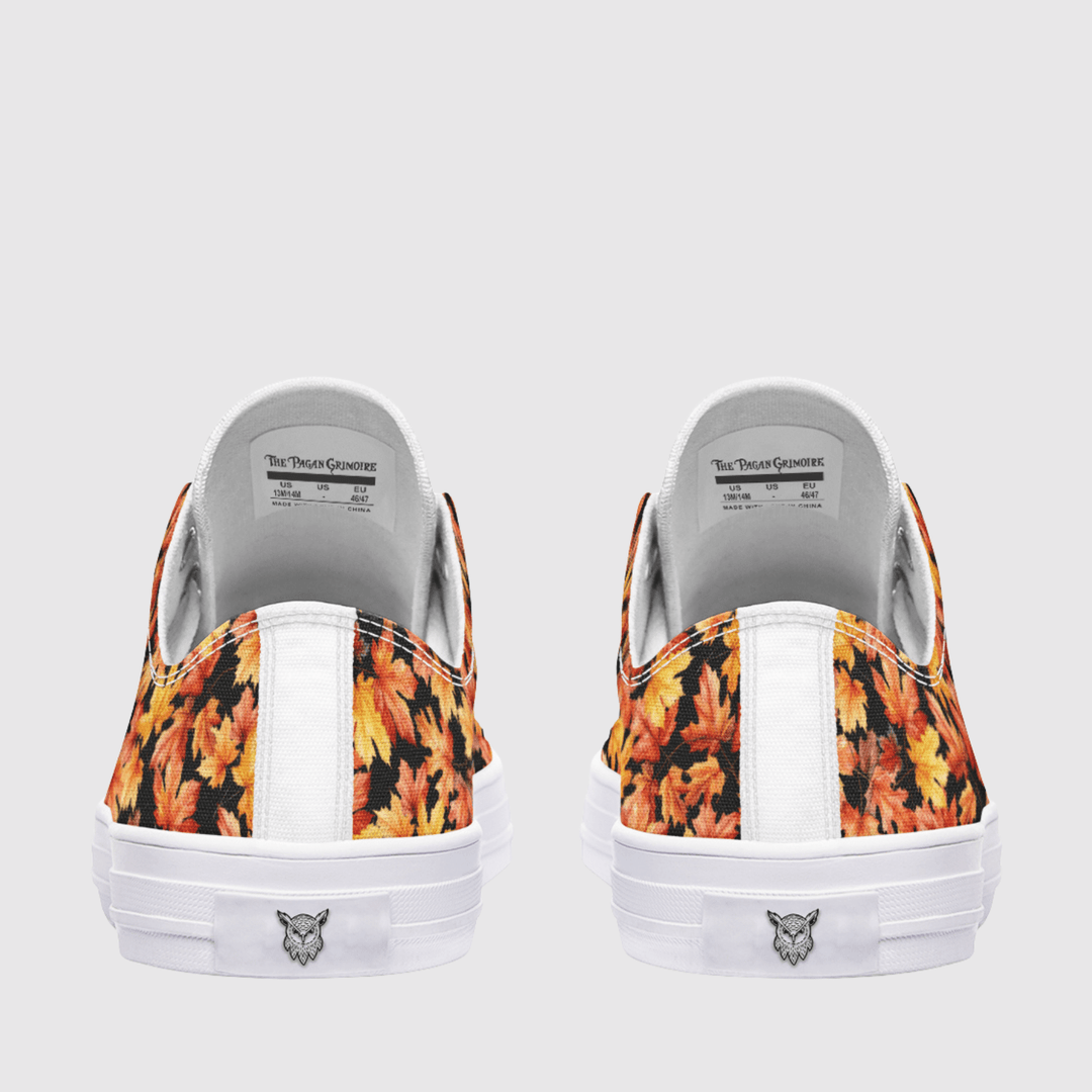 A pair of low-top canvas sneakers shown from the back, featuring a vibrant orange and yellow autumn leaf pattern on the fabric panels, a white vertical stripe, and a white rubber sole with a smalllogo.