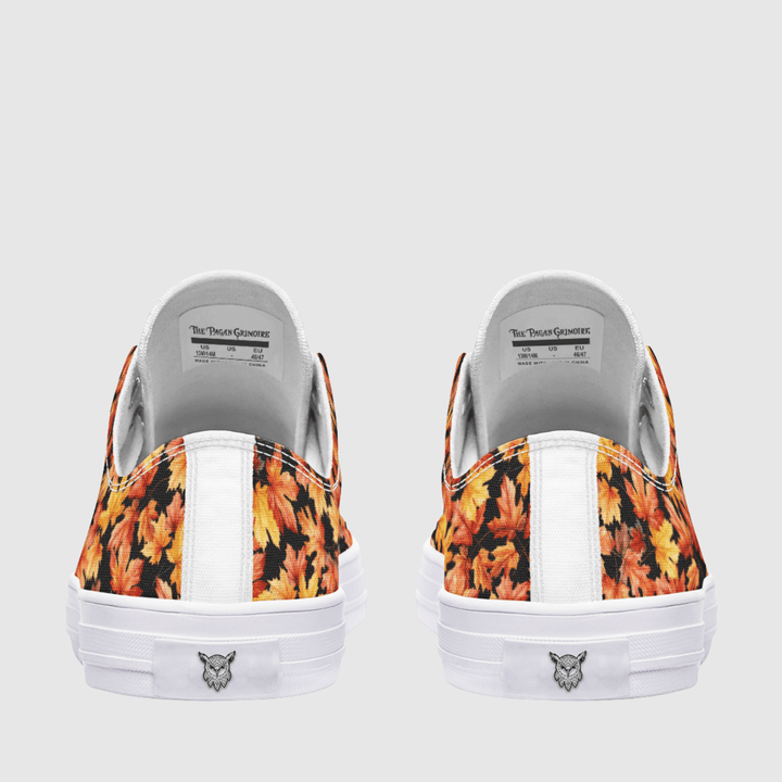 A pair of low-top canvas sneakers shown from the back, featuring a vibrant orange and yellow autumn leaf pattern on the fabric panels, a white vertical stripe, and a white rubber sole with a smalllogo.