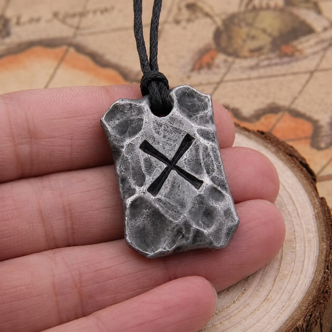 A close-up of a hand holding a textured gray stone pendant necklace with a black engraved symbol of the Gebo rune, suspended from a black cord, with a vintage map and wood slice background.