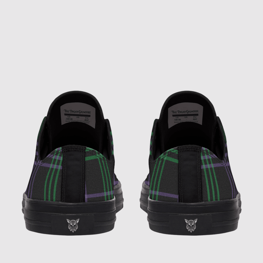 A pair of low-top slip-on sneakers shown from the back, featuring a black base with a green and purple tartan plaid pattern on the sides and a black sole with a small silver cat logo.