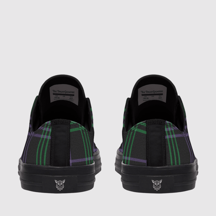 A pair of low-top slip-on sneakers shown from the back, featuring a black base with a green and purple tartan plaid pattern on the sides and a black sole with a small silver cat logo.