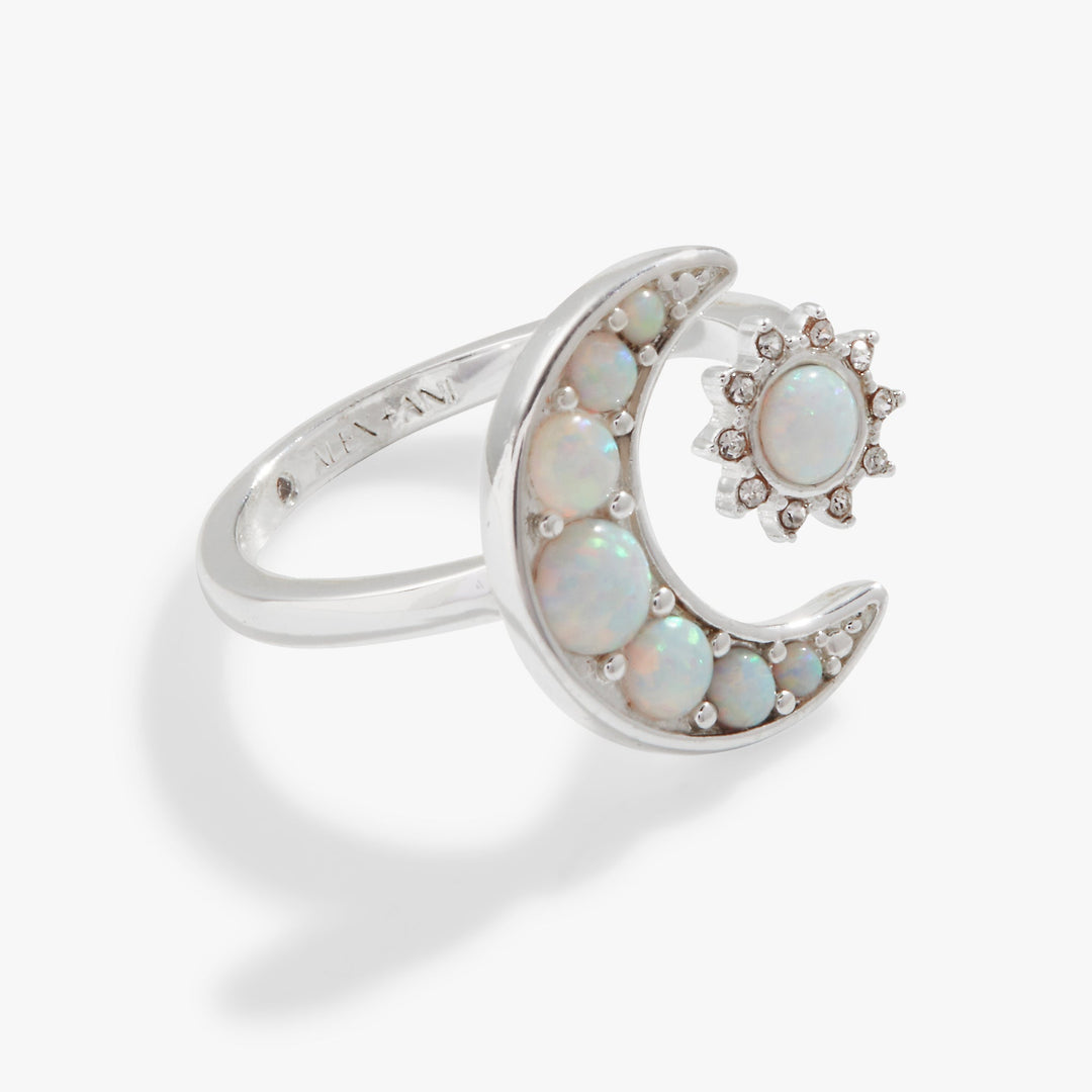 A silver ring featuring a crescent moon and sunburst design, both adorned with small iridescent white opal stones and accented by tiny clear gemstones, shown on a light gray background.