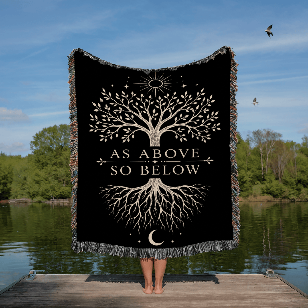 Person holding a black blanket with a tree design and 'As Above So Below' text by a lake.