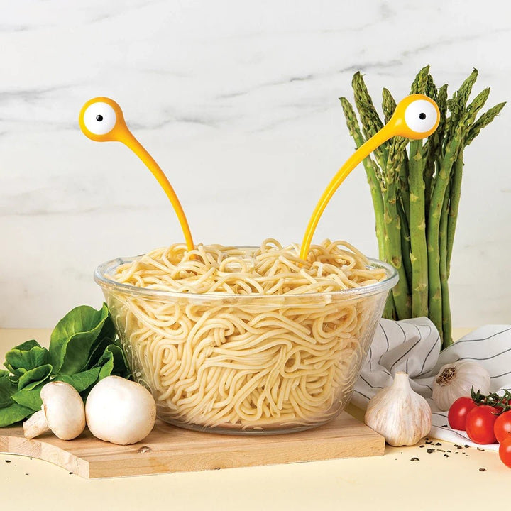 Bowl of spaghetti with monster-like yellow arms reaching into it, surrounded by vegetables on a wooden board.