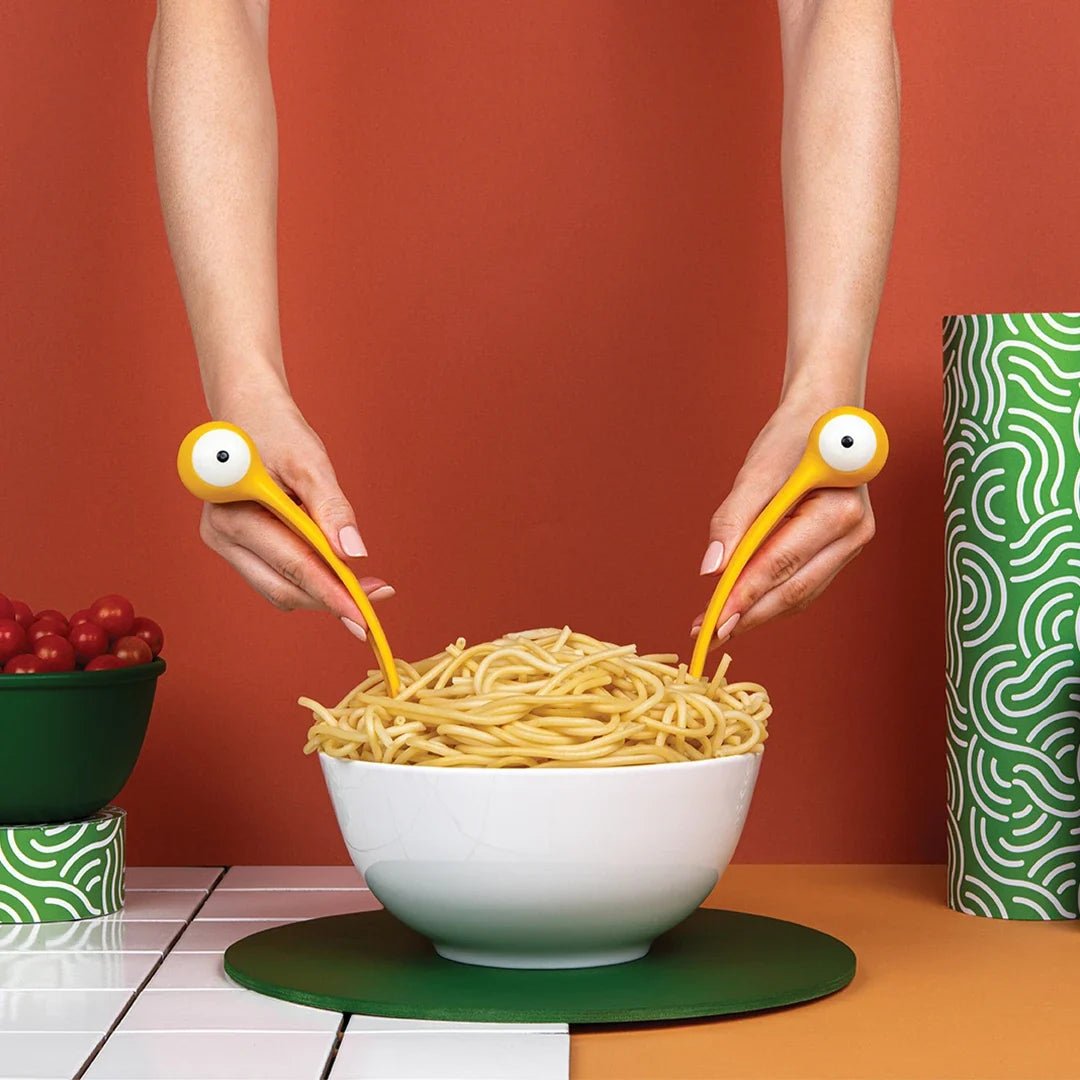 Person using yellow utensils with googly eyes to eat spaghetti from a white bowl on a red background.