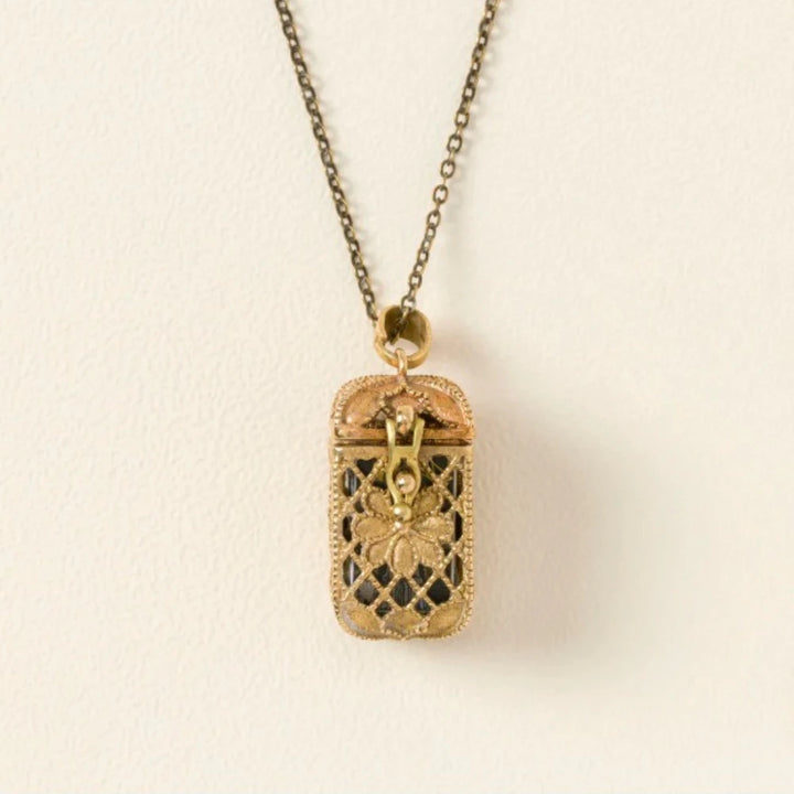 A vintage-style rectangular locket pendant necklace with a gold-tone chain and a box-shaped locket featuring a floral cutout design and black enamel details, shown against a plain off-white background.