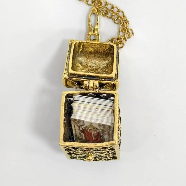 A gold-toned necklace pendant shaped like an open treasure chest, with a short chain visible at the top and a small stack of illustrated cards or pages inside the chest, shown on a plain light background.