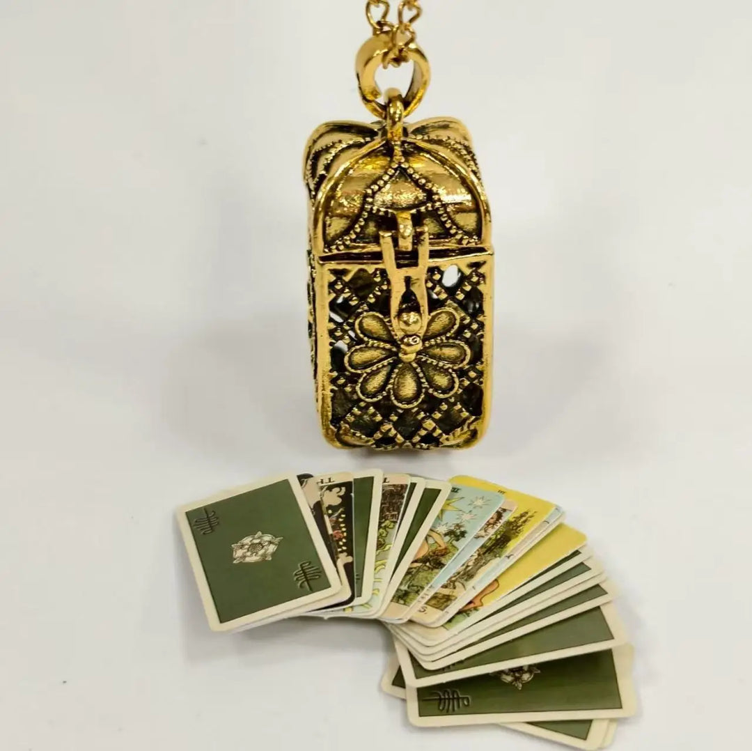 A gold-toned ornate pendant box with a hanging chain, displayed above a fan of illustrated tarot cards on a plain white background.