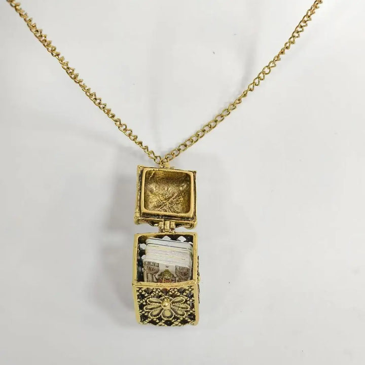 A gold chain necklace with a box-shaped locket pendant, shown open against a plain white background. The locket features a gold-tone floral frame and a small interior containing white and gold elements.