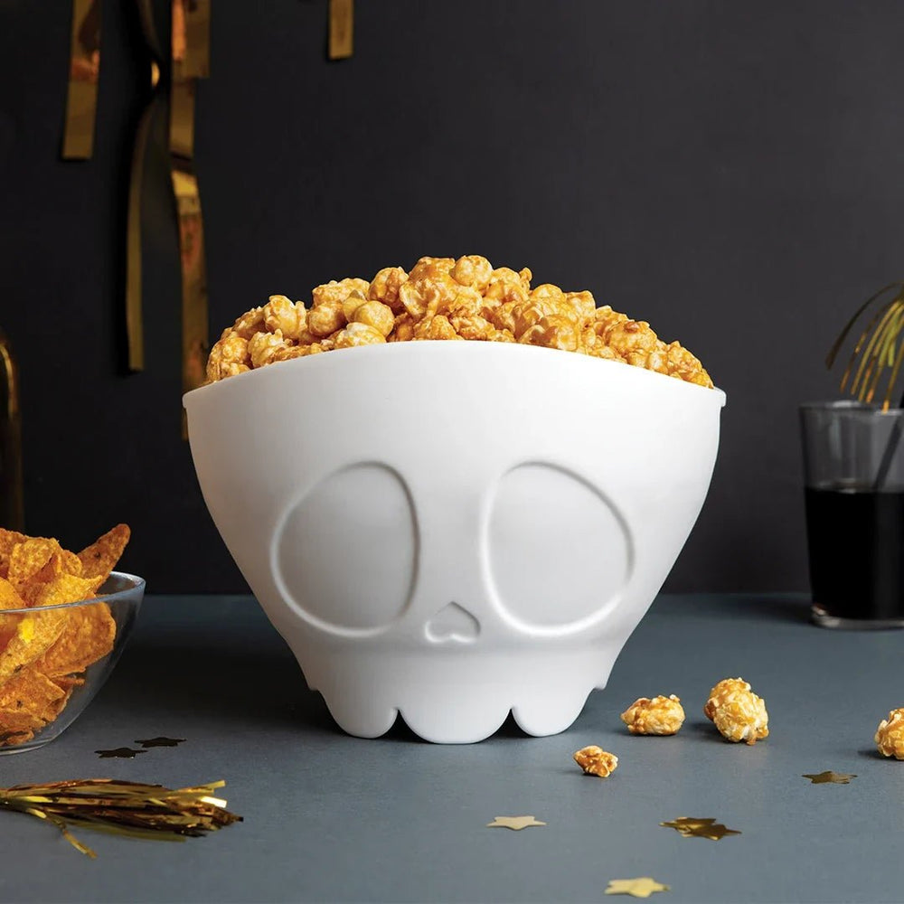 White skull-shaped bowl filled with popcorn on a dark surface with gold popcorn.
