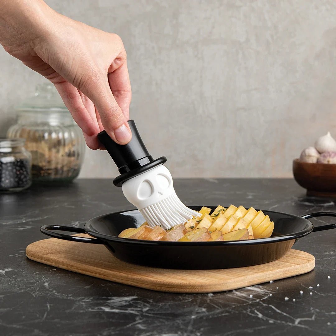 Person using a skull-shaped basting brush to apply sauce to food on a pan.