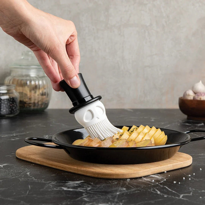 Person using a skull-shaped basting brush to apply sauce to food on a pan.
