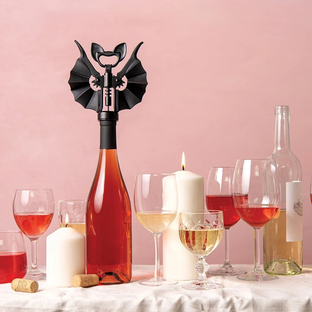 A black winged corkscrew opener tool mounted on a wine bottle, styled on a white tablecloth with wine glasses, white candles, and empty wine bottles against a soft pink background.