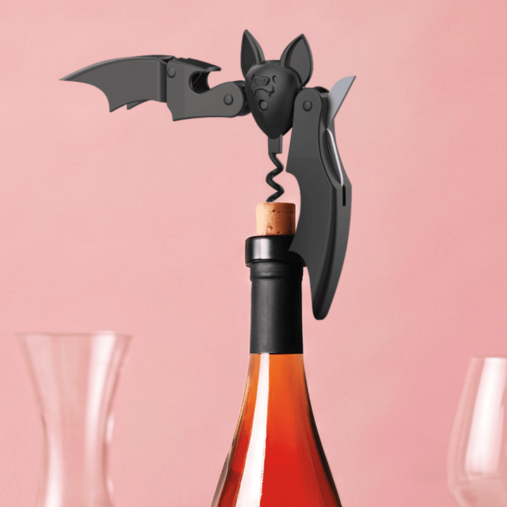 A black bat-shaped corkscrew opener tool with wings and a corkscrew, shown attached to a wine bottle stopper on a pink background with blurred wine glasses at the sides.