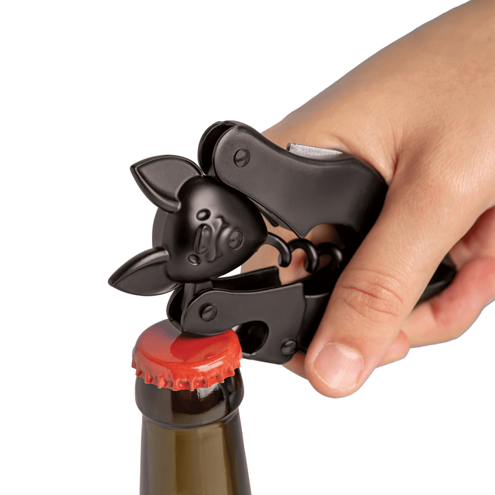 A hand using a black bat-shaped bottle opener to remove a red-capped bottle cap on a white background.