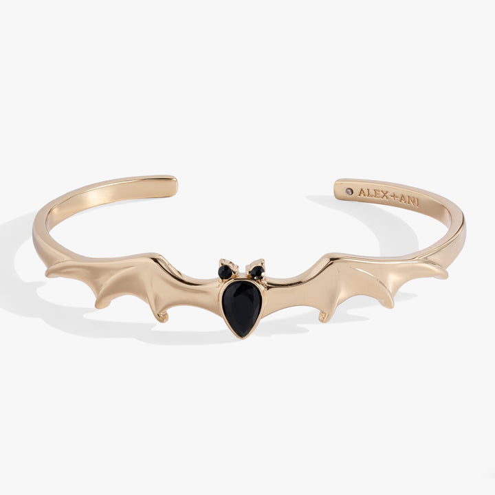 A gold-tone cuff bracelet featuring a bat silhouette with a black teardrop gemstone at the center and two small black round stones for eyes. The inside of the band is engraved with the brand name 'ALEX+ANI'.