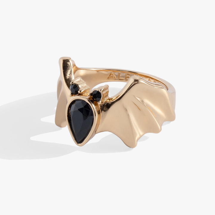 A gold ring shaped like a bat wing, featuring a large teardrop-shaped black gemstone in the center and two small black round stones near the top, shown on a light gray background.