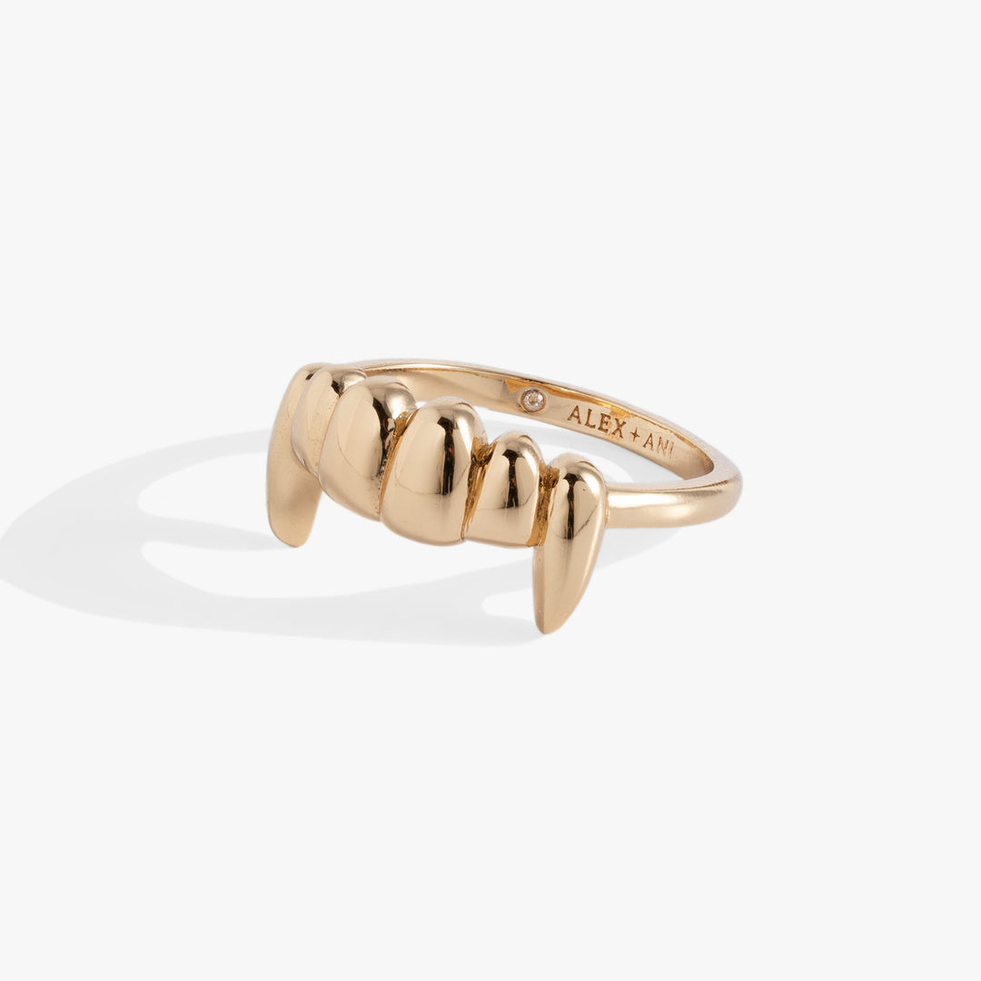 A gold ring shaped like vampire fangs, shown on a clean white background with a soft shadow. The inner band is engraved with the brand name 'ALEX+AND'.